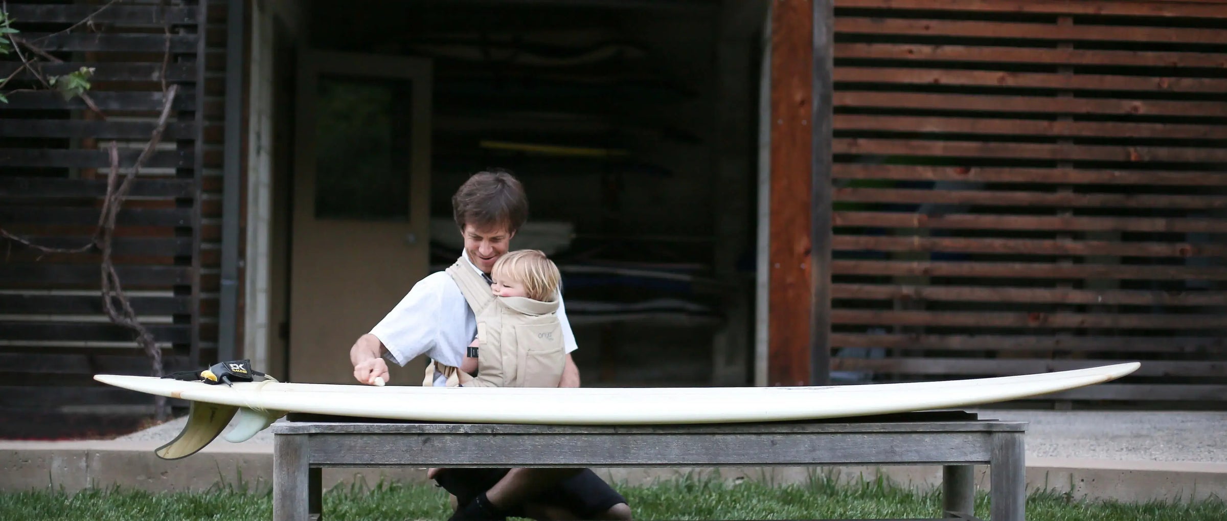 Man waxing a surfboard while wearing baby in front carry position