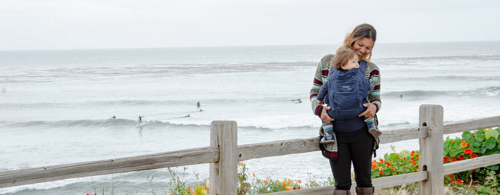 Smiling woman overlooking the ocean while carrying baby in an Onya Baby Cruiser in the front carry position