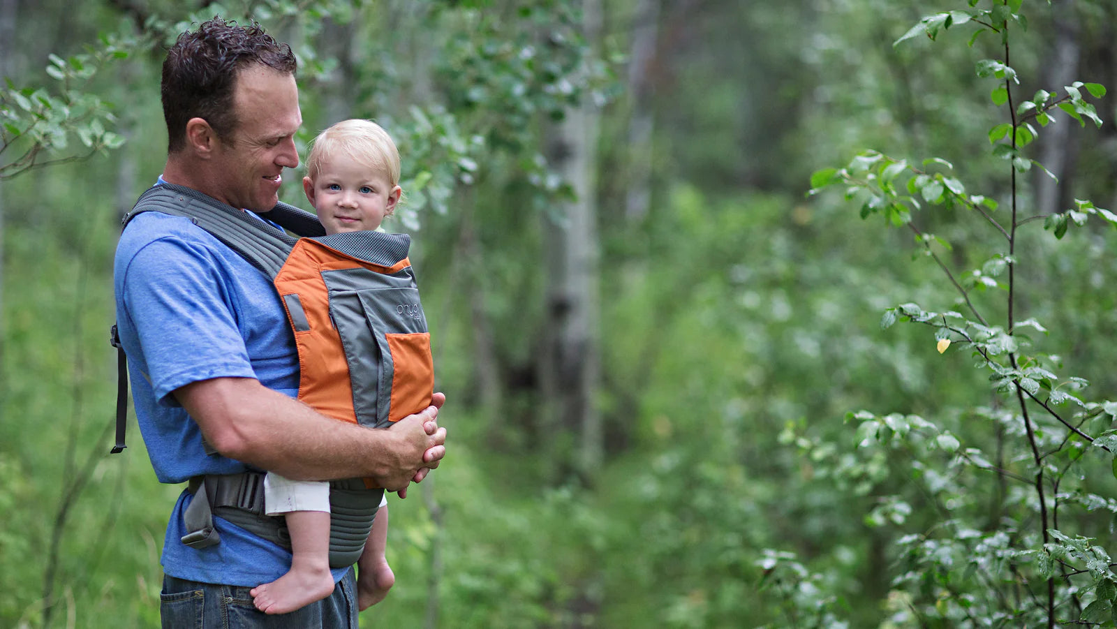 Man carrying smiling child in an Onya Baby Outback in front carry position while walking on a forest trail