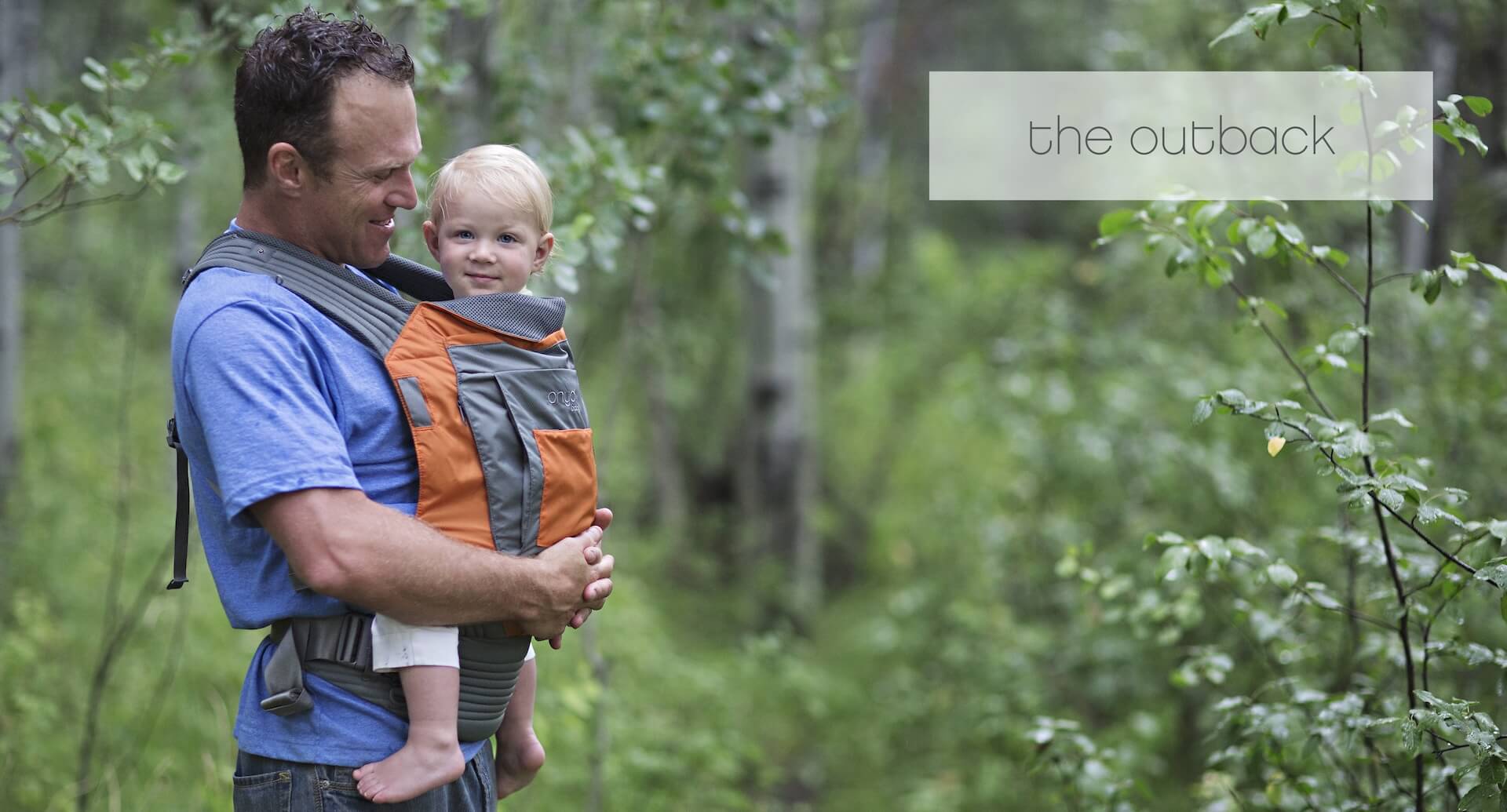 Man carrying smiling child in an Onya Baby Outback in front carry position while walking on a forest trail