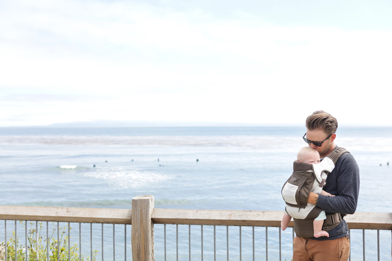 Man standing overlooking a beach with baby in an Onya Baby Outback in the front carry position