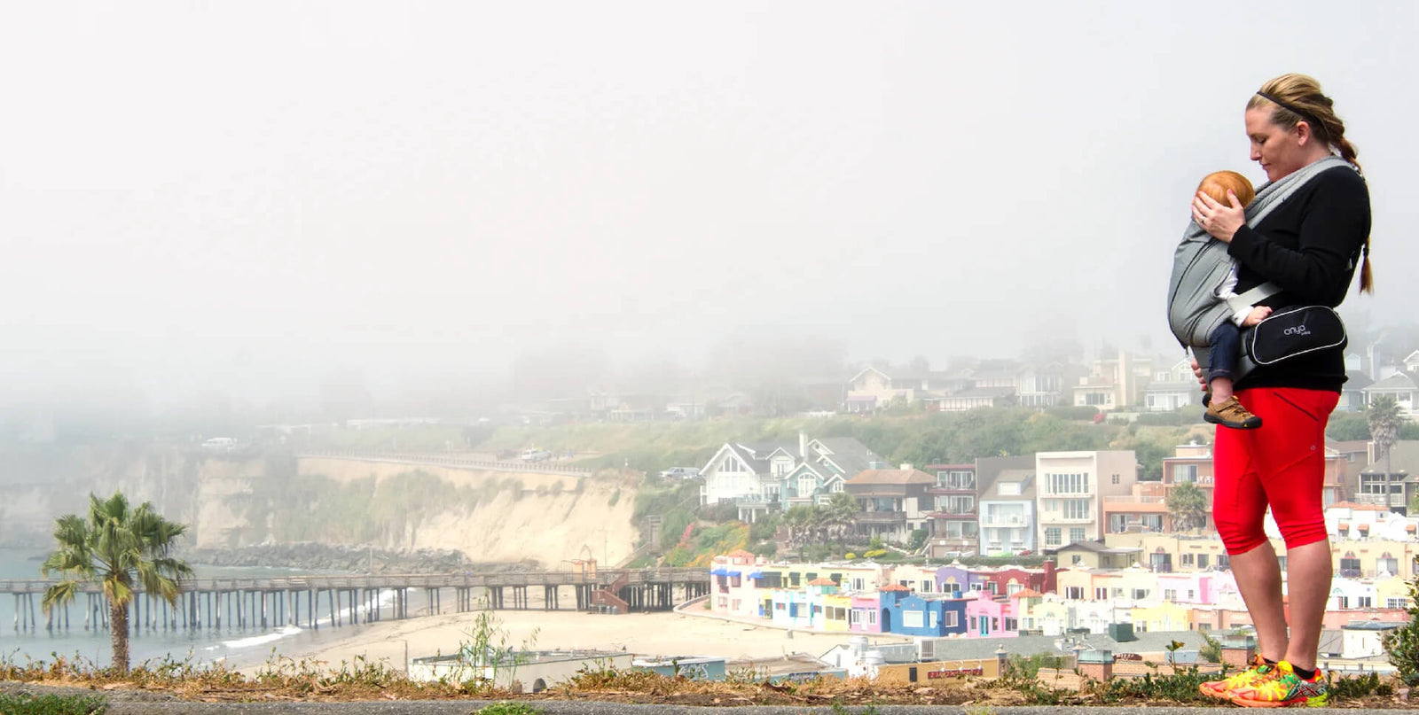 Side View of woman standing overlooking a beach with baby in an Onya Baby Pure in the front carry position wearing the Hipster Bag
